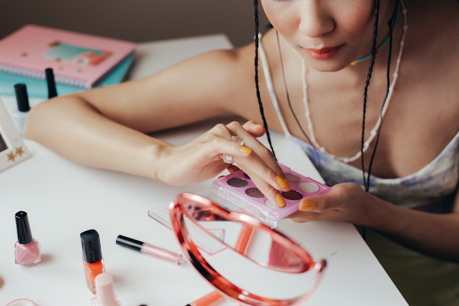 Woman using eyeshadow palette with cosmetics scattered on a table, reflecting in mirror.
