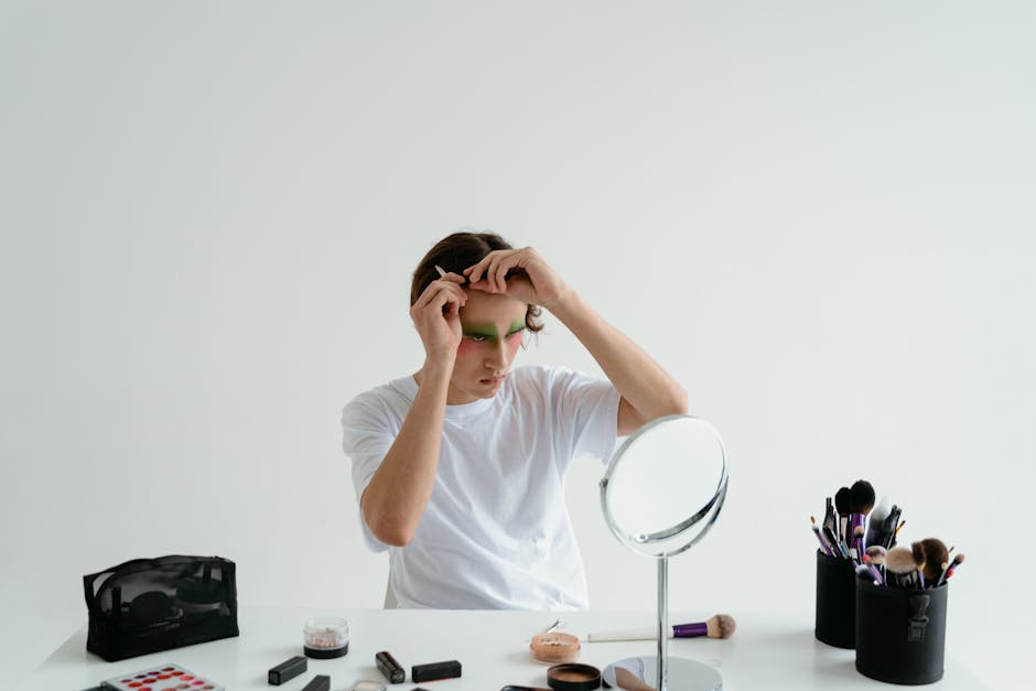 A young man in a white shirt applying makeup in front of a mirror, surrounded by various cosmetic tools