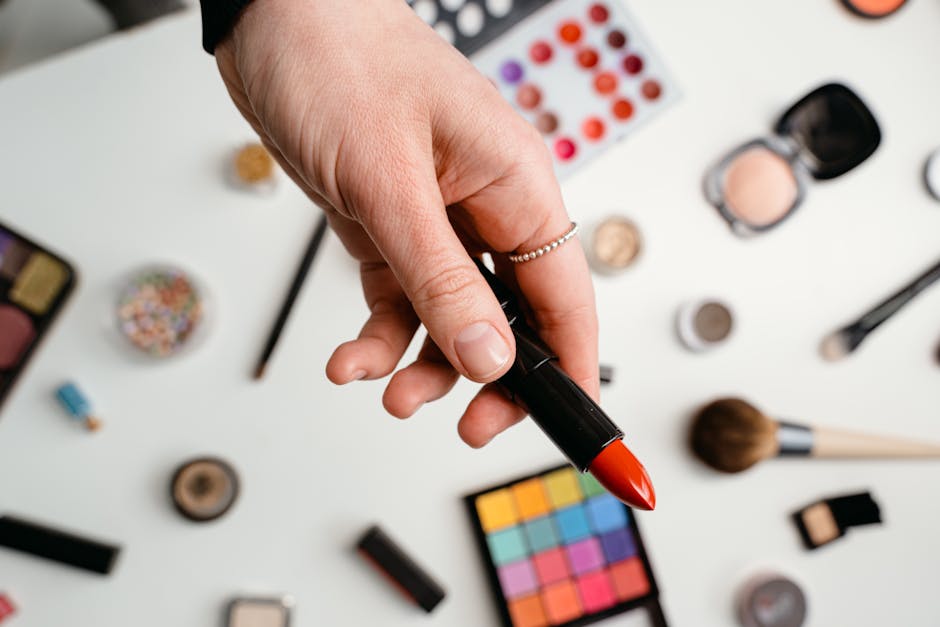 A hand holding a red lipstick surrounded by various makeup products on a light background