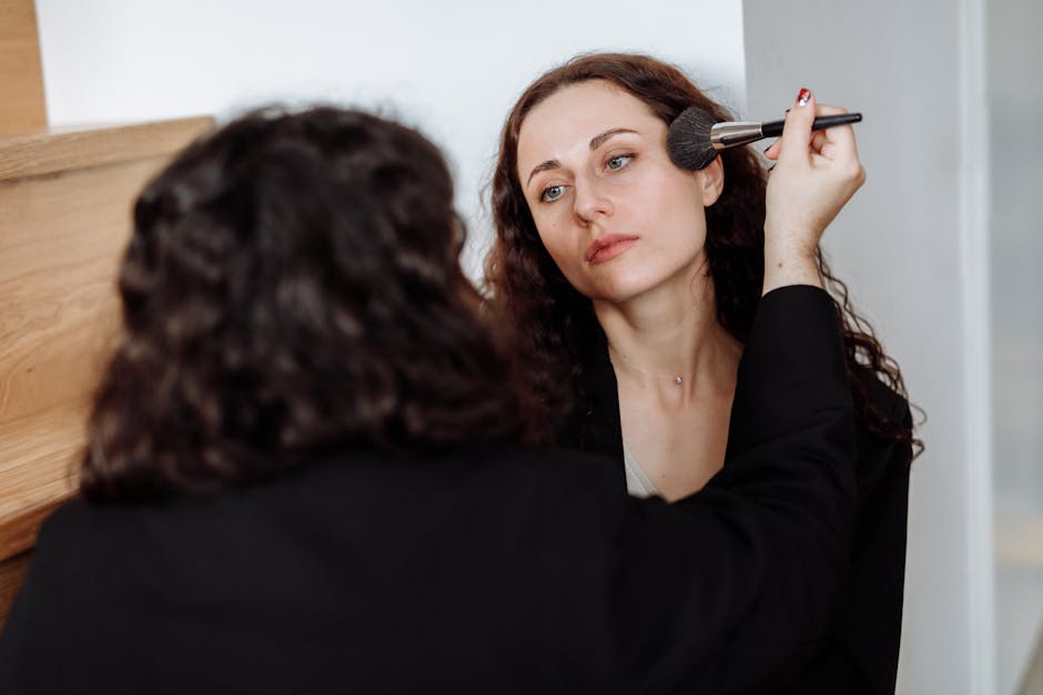 A woman uses a makeup brush to apply cosmetics to another woman in a cozy indoor setting.